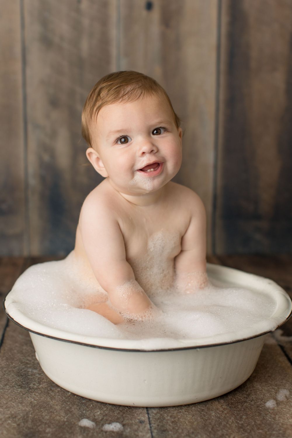 Happy baby playing in bubble bath in white basin against rustic wooden background.