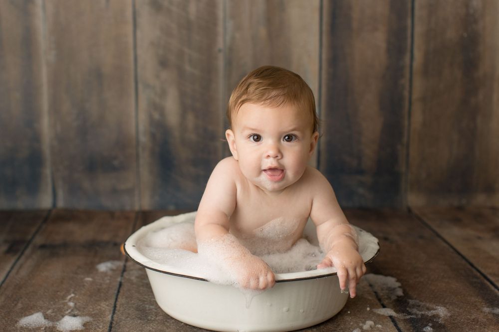 Baby sitting in white wash basin with soap bubbles against wooden wall.