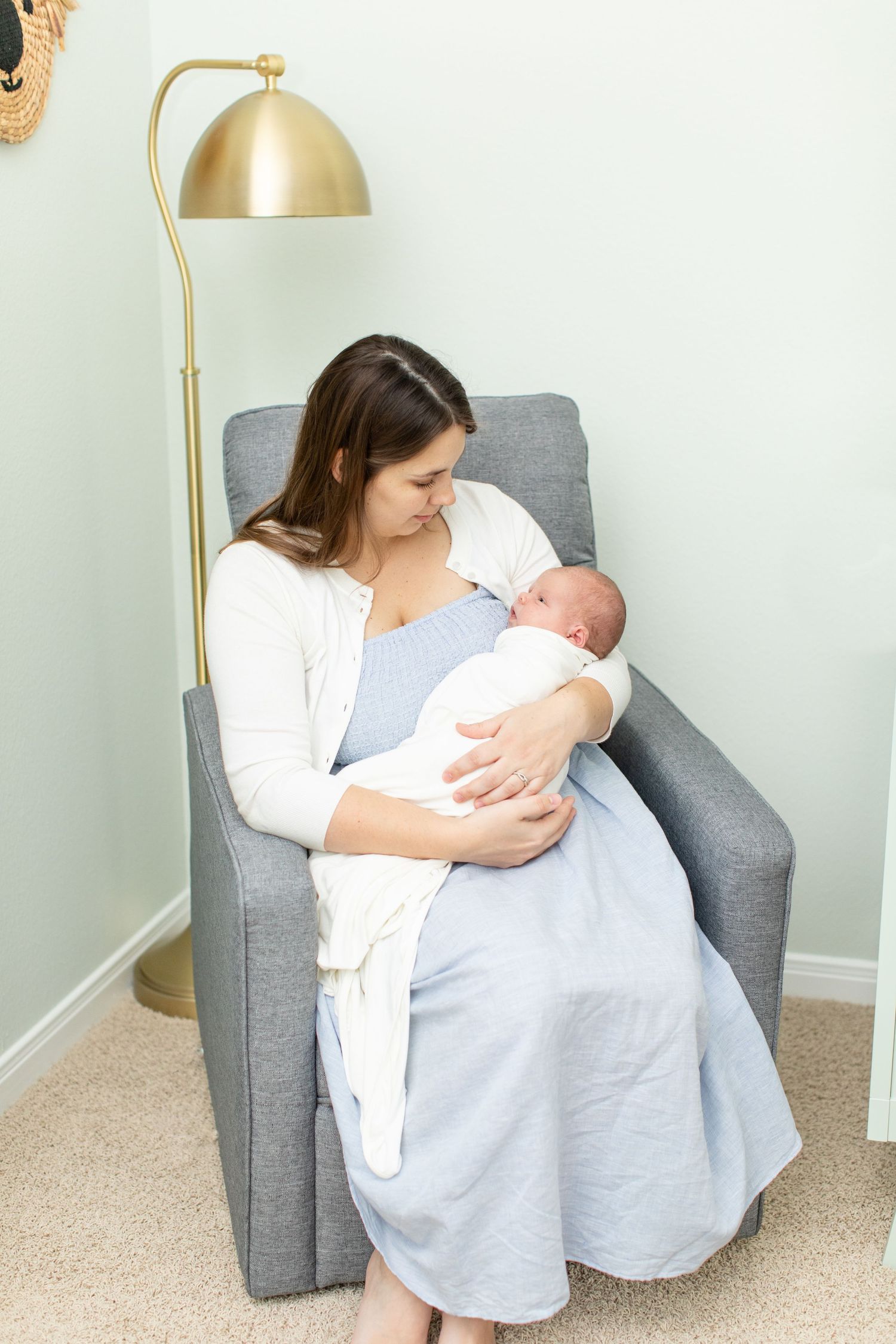 Adult cradles newborn while sitting in grey armchair beneath gold lamp in nursery.
