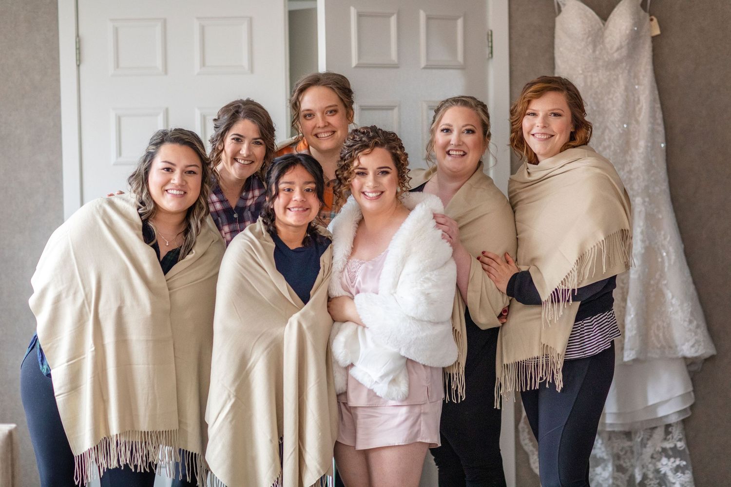A group wearing cozy beige shawls pose together during wedding preparations with a dress visible in background.
