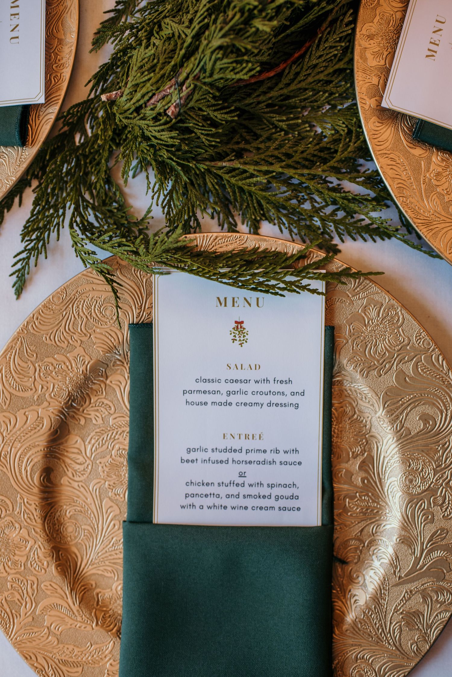 Wedding menu cards with evergreen sprigs placed on textured gold charger plates.