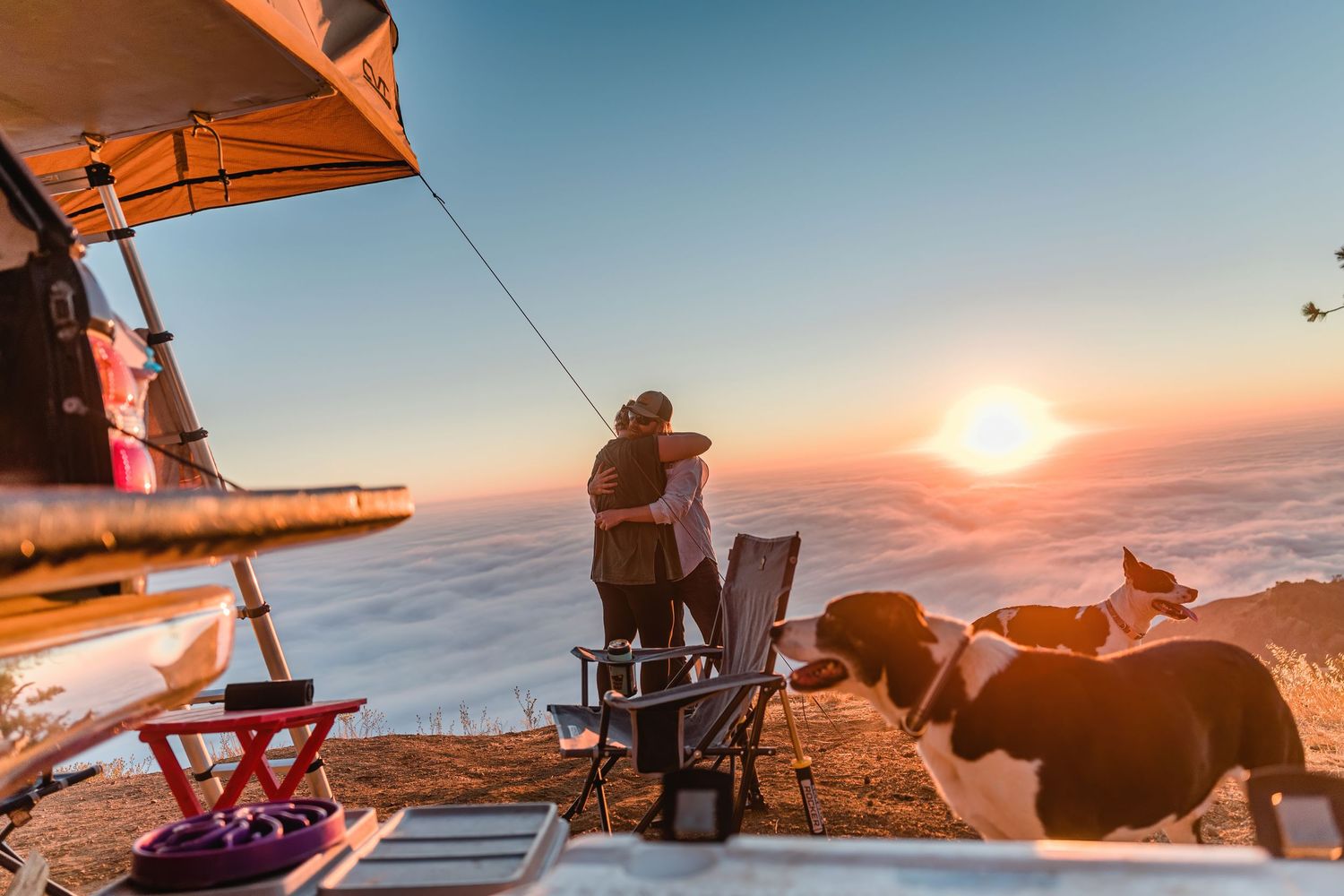 A couple and their dog watch the sunset from a mountaintop campsite with an orange tent.