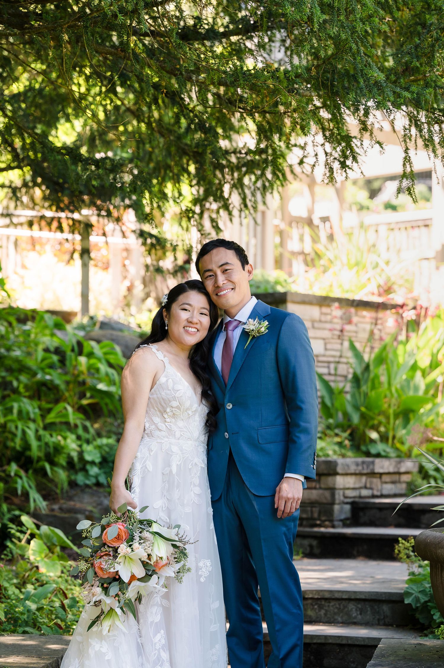 A wedding couple poses on stone garden steps with overhanging trees and flowers.