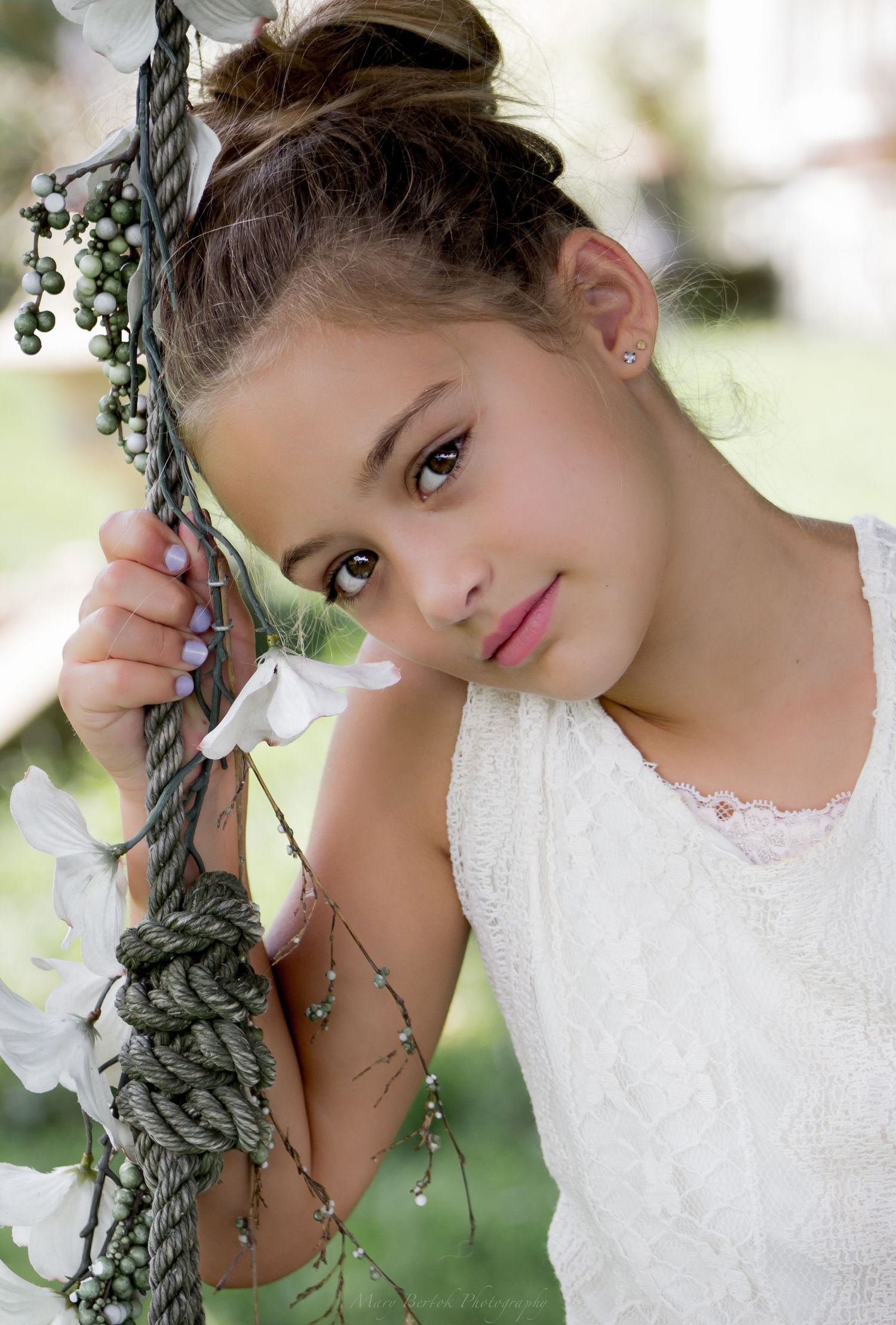 Young girl in white lace dress holds decorative rope with flowers in outdoor portrait.