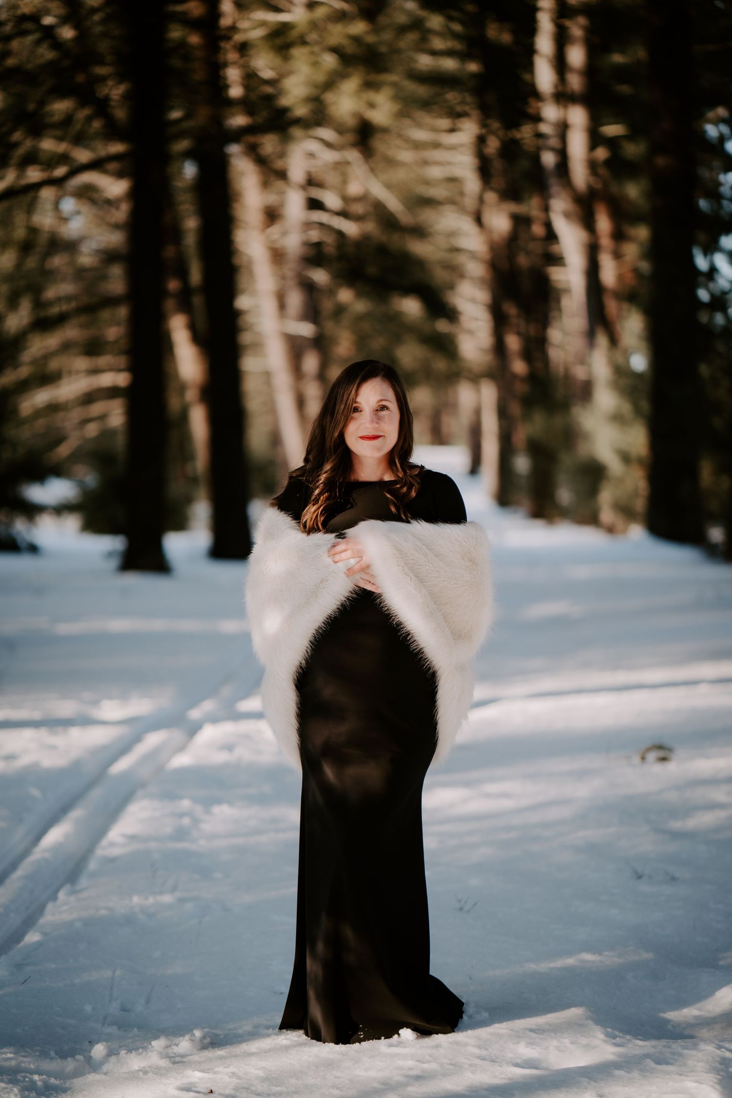 Pregnant mother standing in in the snow in a black dress and a white shawl between a line of trees