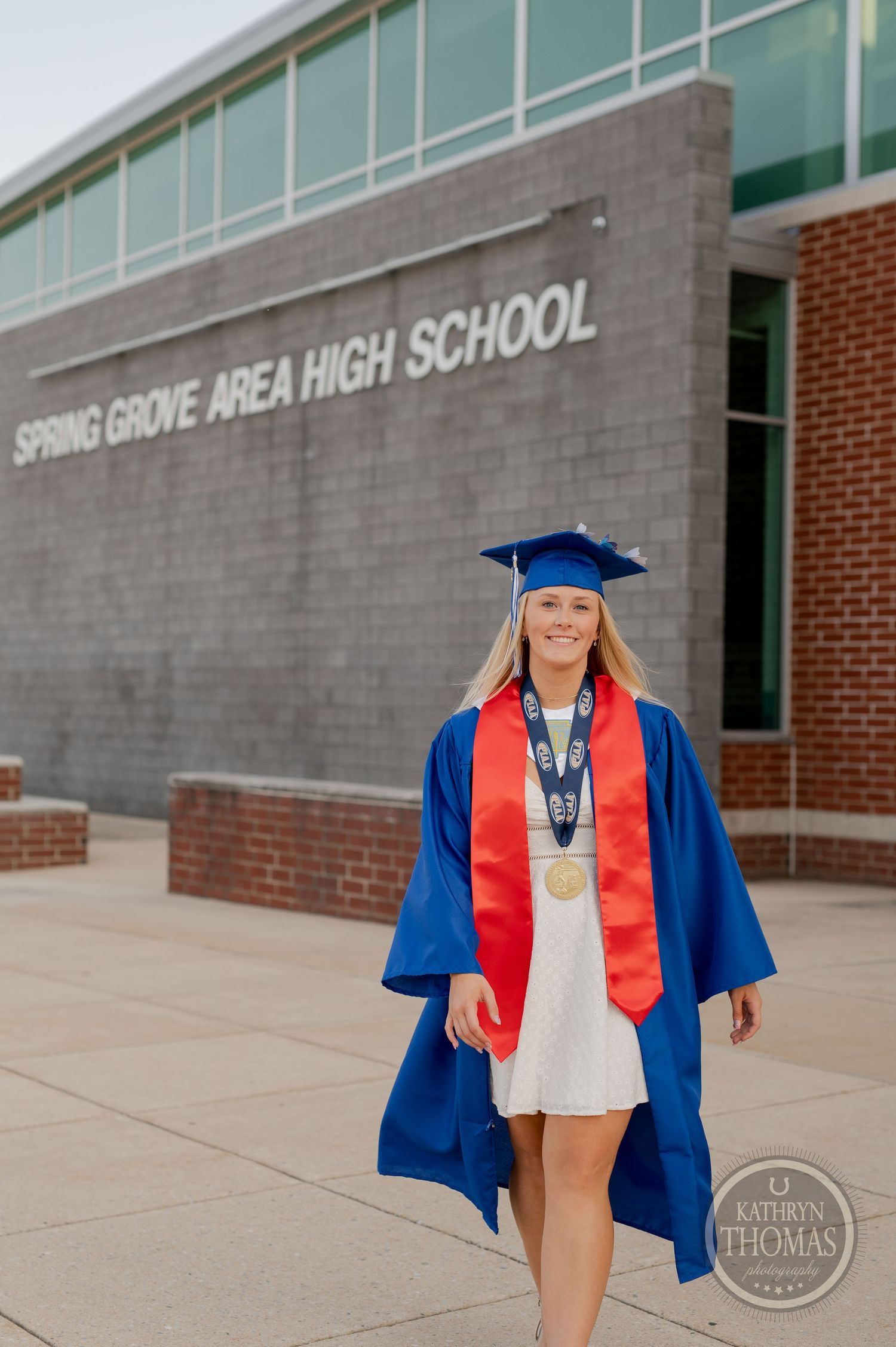 Graduate in blue and red graduation regalia smiles outside high school building.