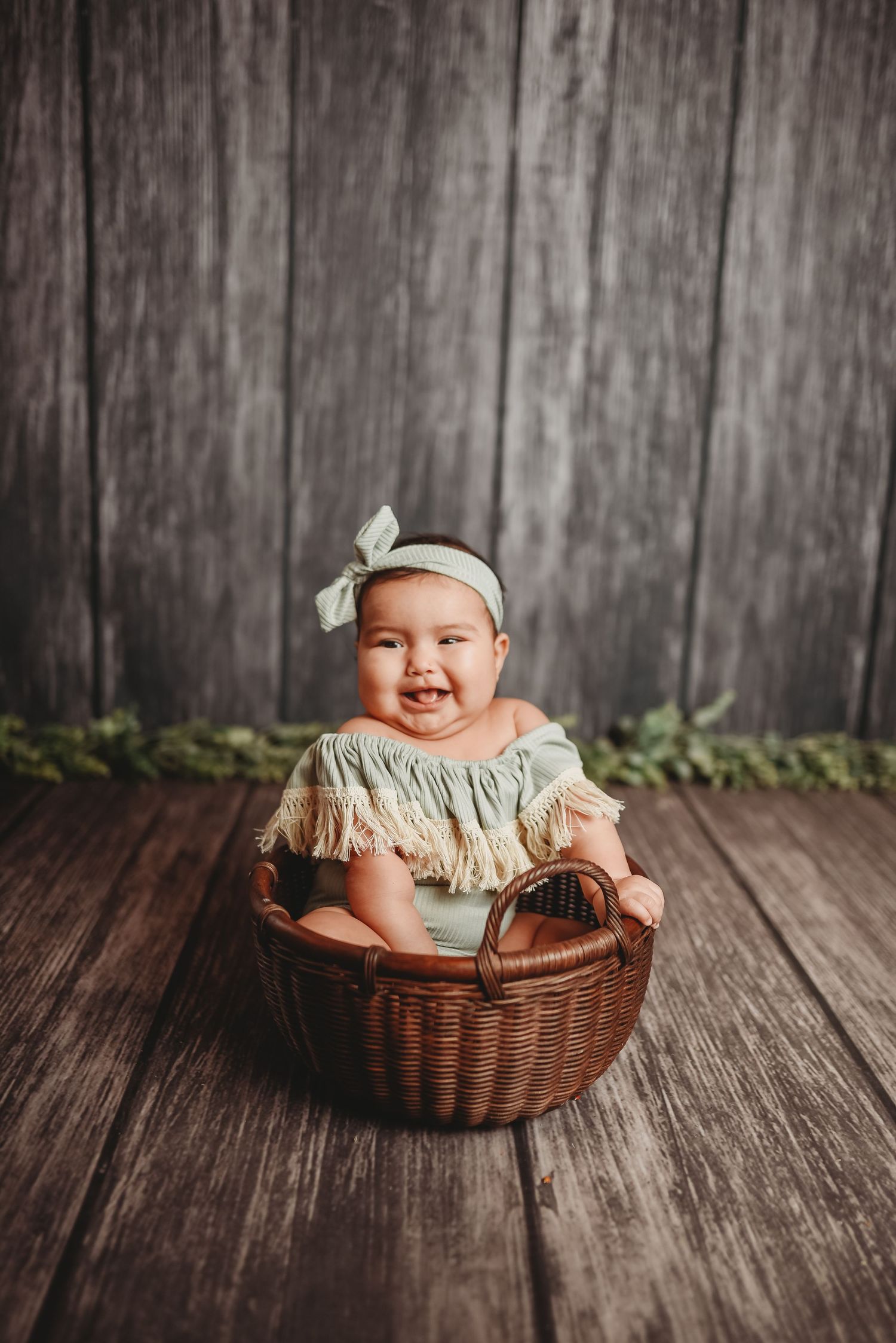 Baby in fringe dress sits in wicker basket against rustic wooden backdrop.