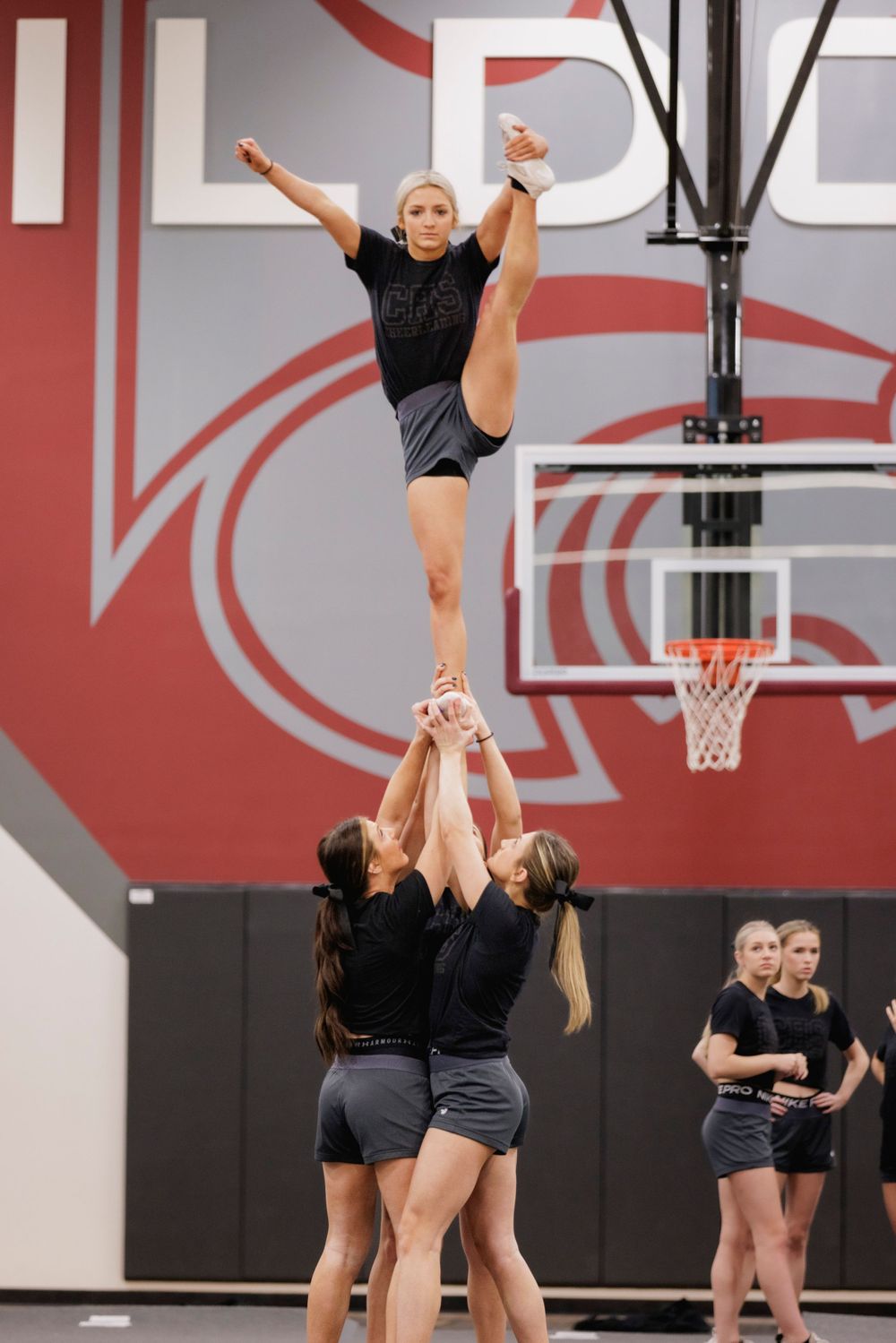 Cheerleaders performing advanced stunt lift in gymnasium with red wall and basketball hoop.