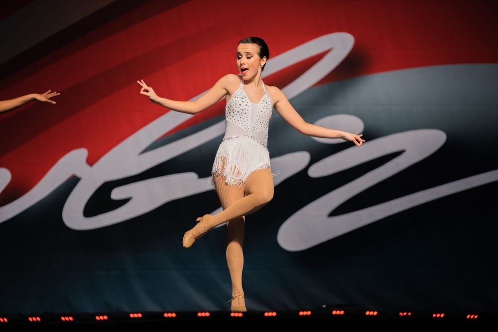 Dancer in white leotard leaps gracefully against red and black backdrop with artistic swirls.