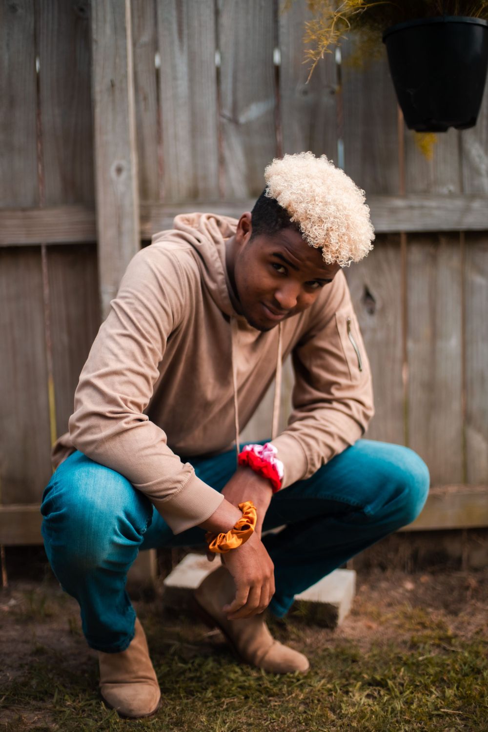 A man in a beige hoodie and blue jeans crouches down against a wooden fence background.