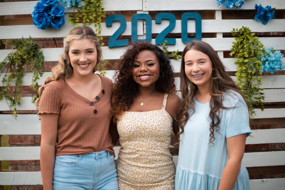 Three young women pose together in front of a decorated wall with 2020 signage and blue flowers.