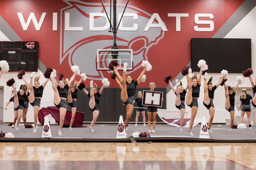 Cheerleaders practicing routine in gym with Wildcats logo and basketball hoop visible.