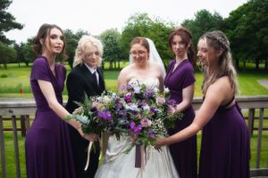 Wedding party in purple dresses admires bride's stunning floral bouquet on outdoor deck.