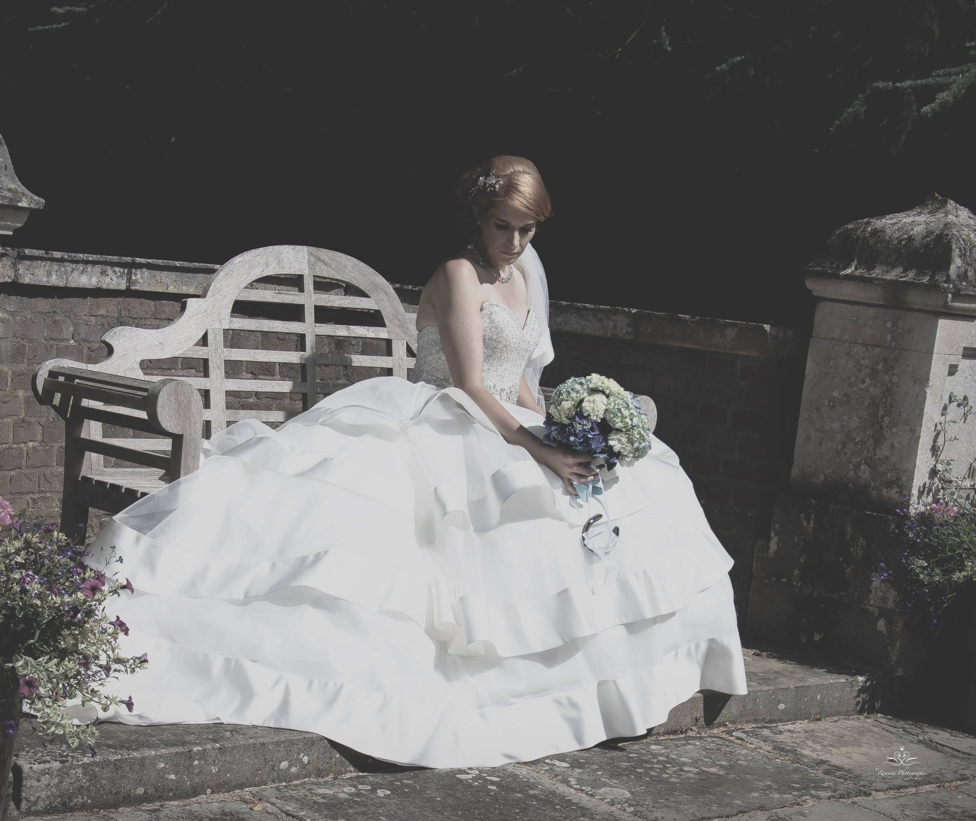 A bride in a white ballgown sits contemplatively on a garden bench holding a bouquet.