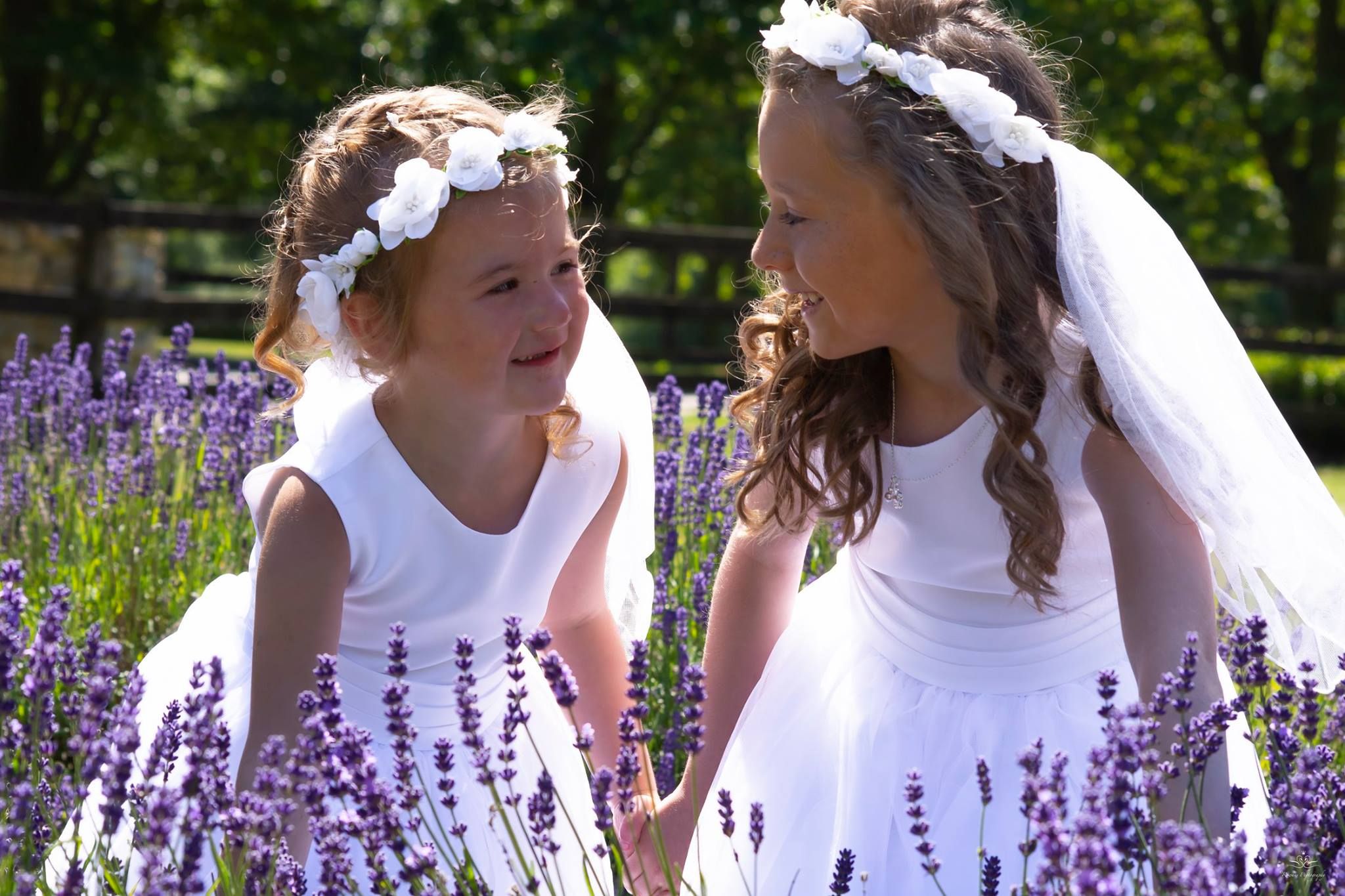 Two flower girls in white dresses and floral crowns share a joyful moment among purple lavender flowers.