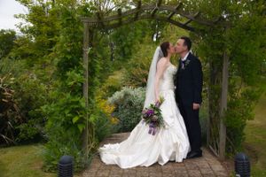 Newlyweds share a romantic moment under a garden archway with purple bouquet and flowing white dress.