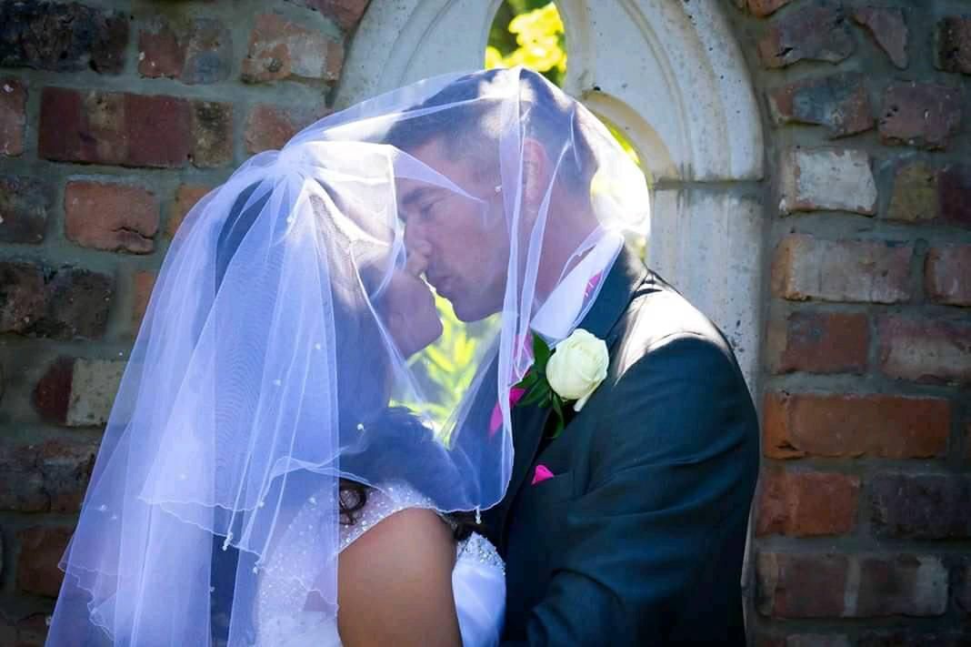 Newlyweds share a romantic kiss under a flowing white veil against a brick wall backdrop.