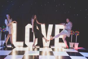 Party guests pose playfully against illuminated LOVE letters on a checkered dance floor.