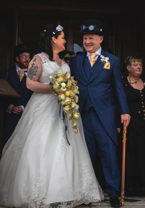 A steampunk-themed wedding couple in blue and white attire pose together with the bride holding a cascading bouquet.