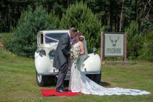 Couple posing by vintage white car at outdoor wedding venue.