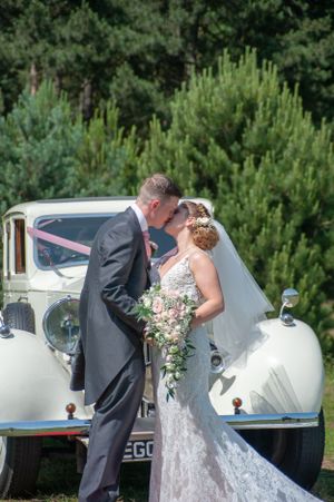 Newlyweds embrace for a romantic kiss by a classic white car surrounded by pine trees.