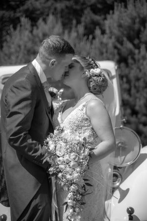 Intimate black and white wedding photo of a couple sharing a tender moment beside a vintage car.