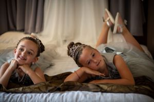 Two young flower girls in tiaras pose playfully on a bed.