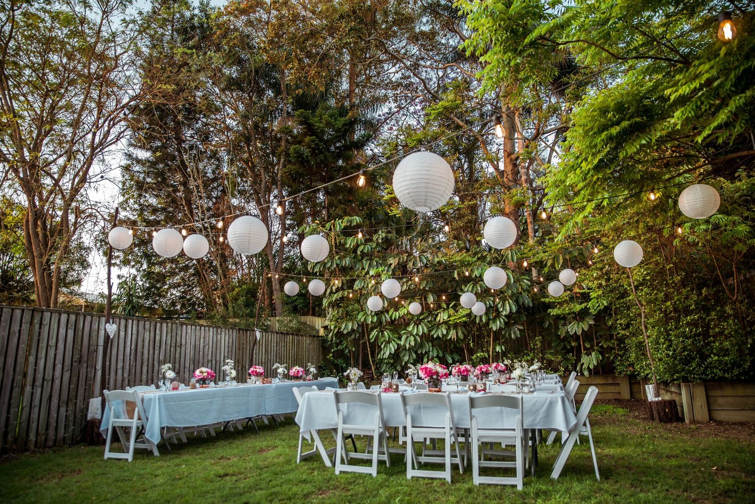 Outdoor wedding reception setup with white paper lanterns hanging in trees at dusk.
