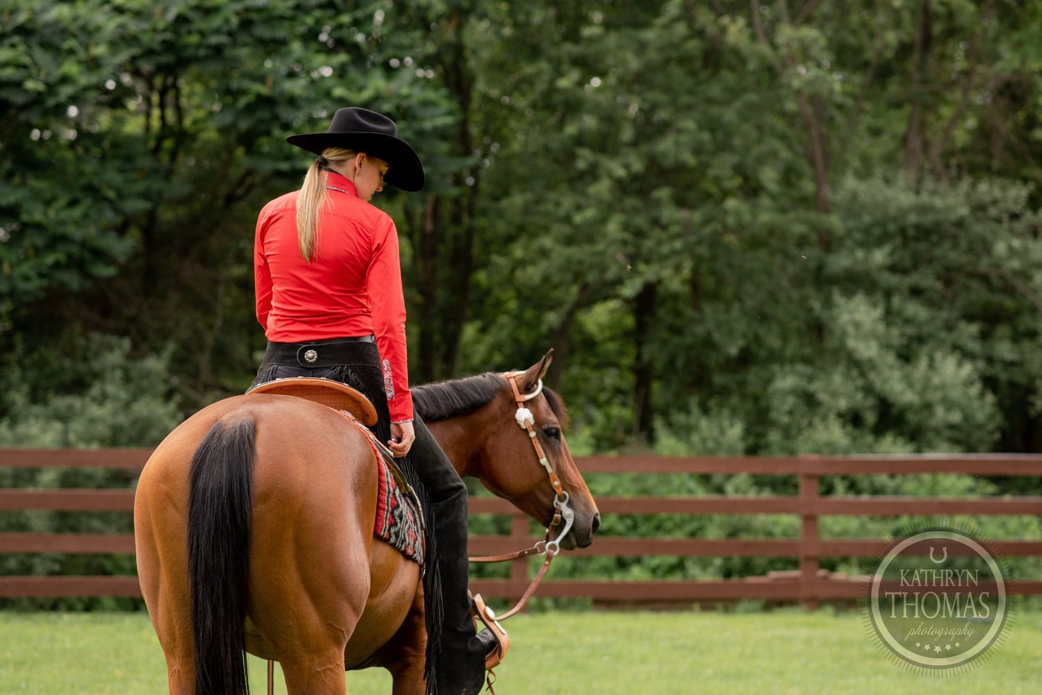 Western rider in red shirt and black hat riding a brown horse in front of wooden fencing.