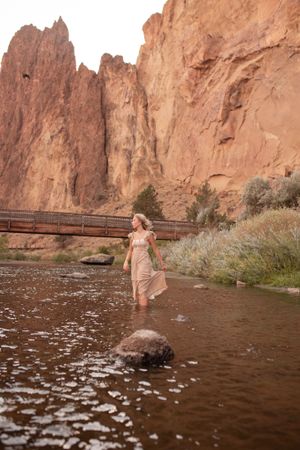 A figure in a flowing dress wading through water near towering red rock formations.