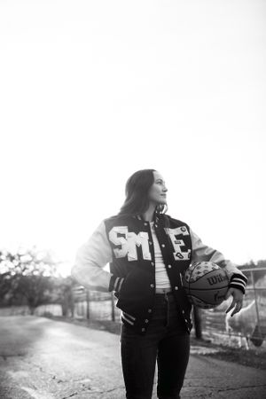 A black and white portrait of someone wearing a varsity jacket and holding a football in an outdoor setting.