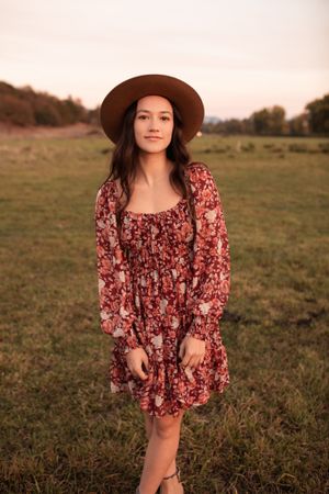 Portrait in a floral dress and brown hat against a sunset meadow backdrop.