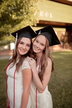 Two graduates in white dresses and black caps pose together on graduation day.