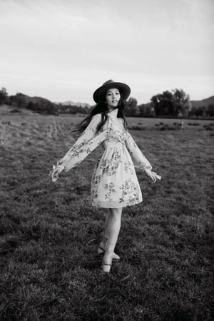 Person in a floral dress and wide-brimmed hat twirling in a meadow captured in black and white.