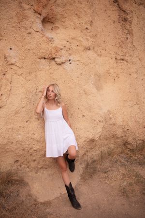 Someone in a white dress and boots poses against an orange rock wall in a desert setting.