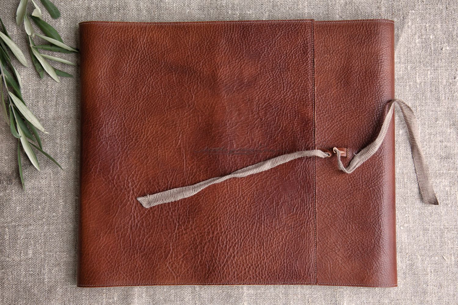 Brown leather photo album or journal with fabric tie closure on linen background with olive branch.