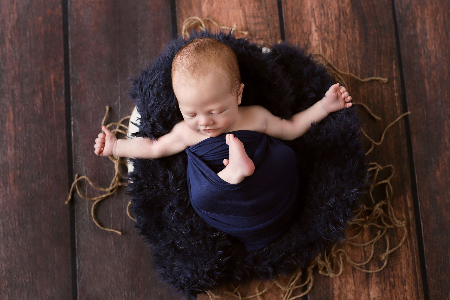Sleeping newborn in navy outfit lying on dark textured surface with arms outstretched.