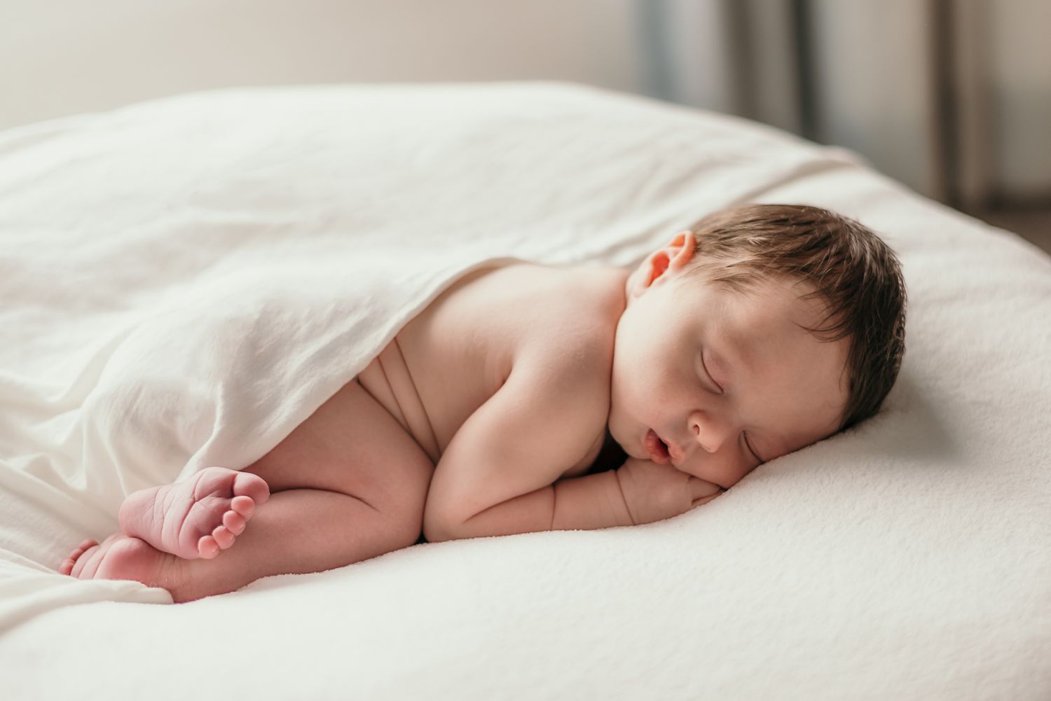Sleeping newborn baby wrapped in white blanket lying on soft white bedding.