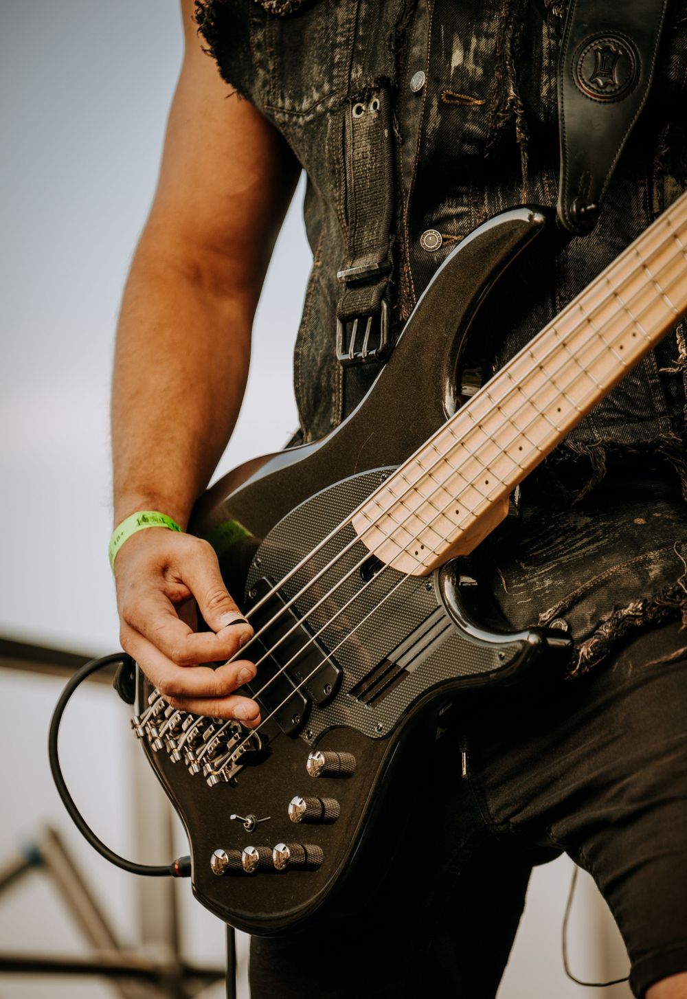 Close-up shot of a black bass guitar being played by hands wearing a leather vest.