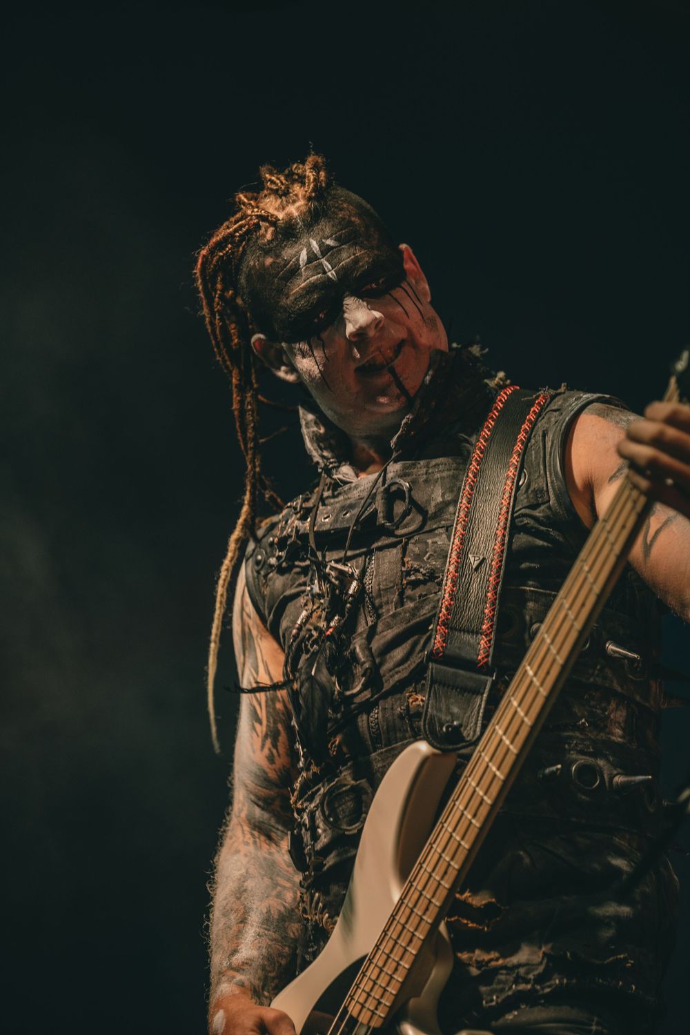 A dramatic performance shot of a bassist with face paint and dreadlocks in dark lighting.