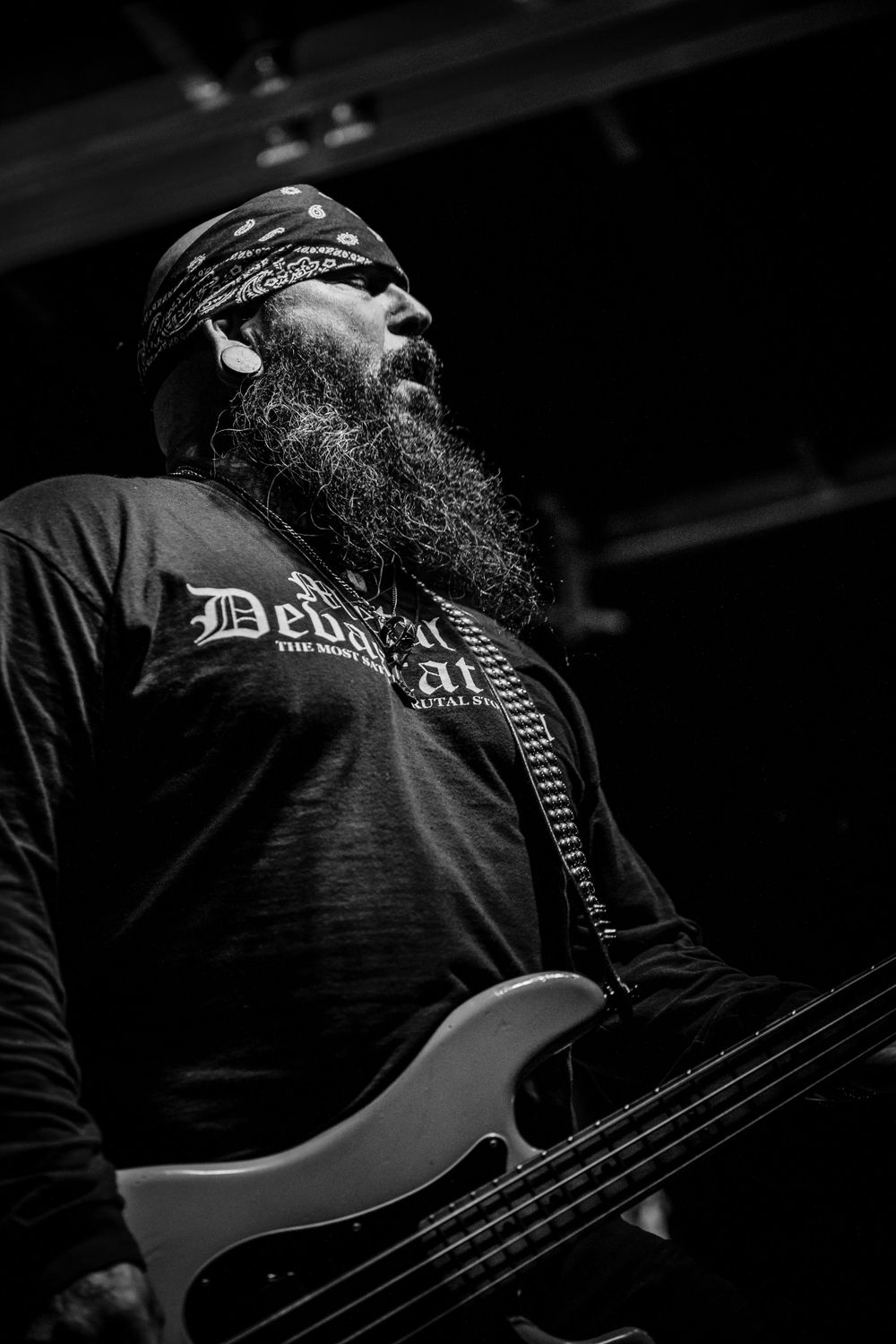 Heavy metal bassist with long beard wearing bandana and band t-shirt performs on stage in black and white.