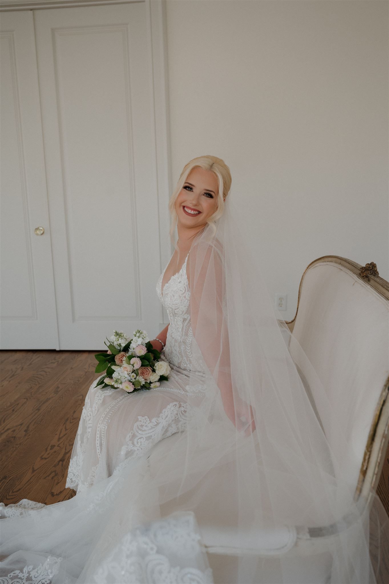A bride in white lace sits on a vintage settee, holding flowers and wearing a flowing veil.
