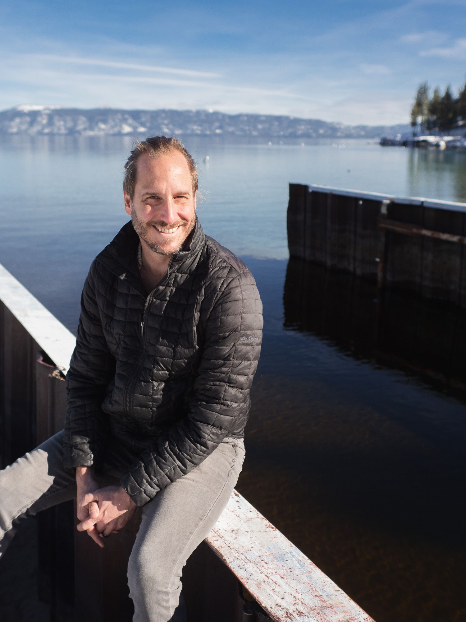 Kris Swanson in a black puffer jacket sits along waterfront railing with snow-capped mountains and calm lake in background.