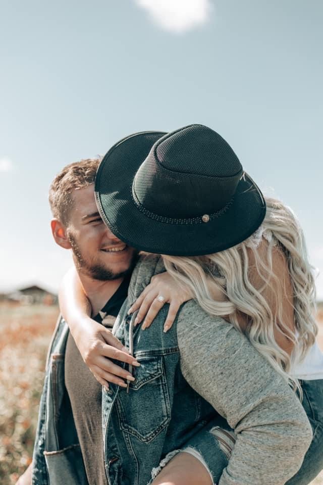 Smiling man giving piggyback ride to blonde woman in black hat in sunny outdoor field.