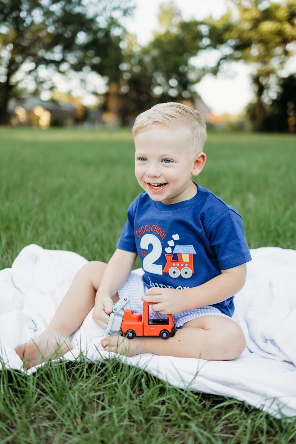 Young child in blue t-shirt plays with toy truck while lying on white blanket in grassy field.