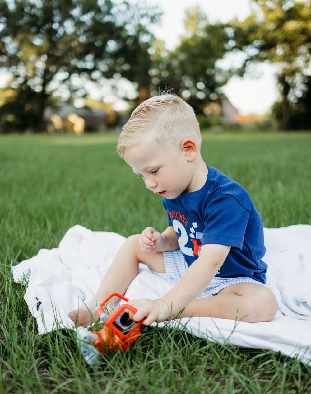 Child lying on white blanket plays with orange toy truck in sunny grass field.