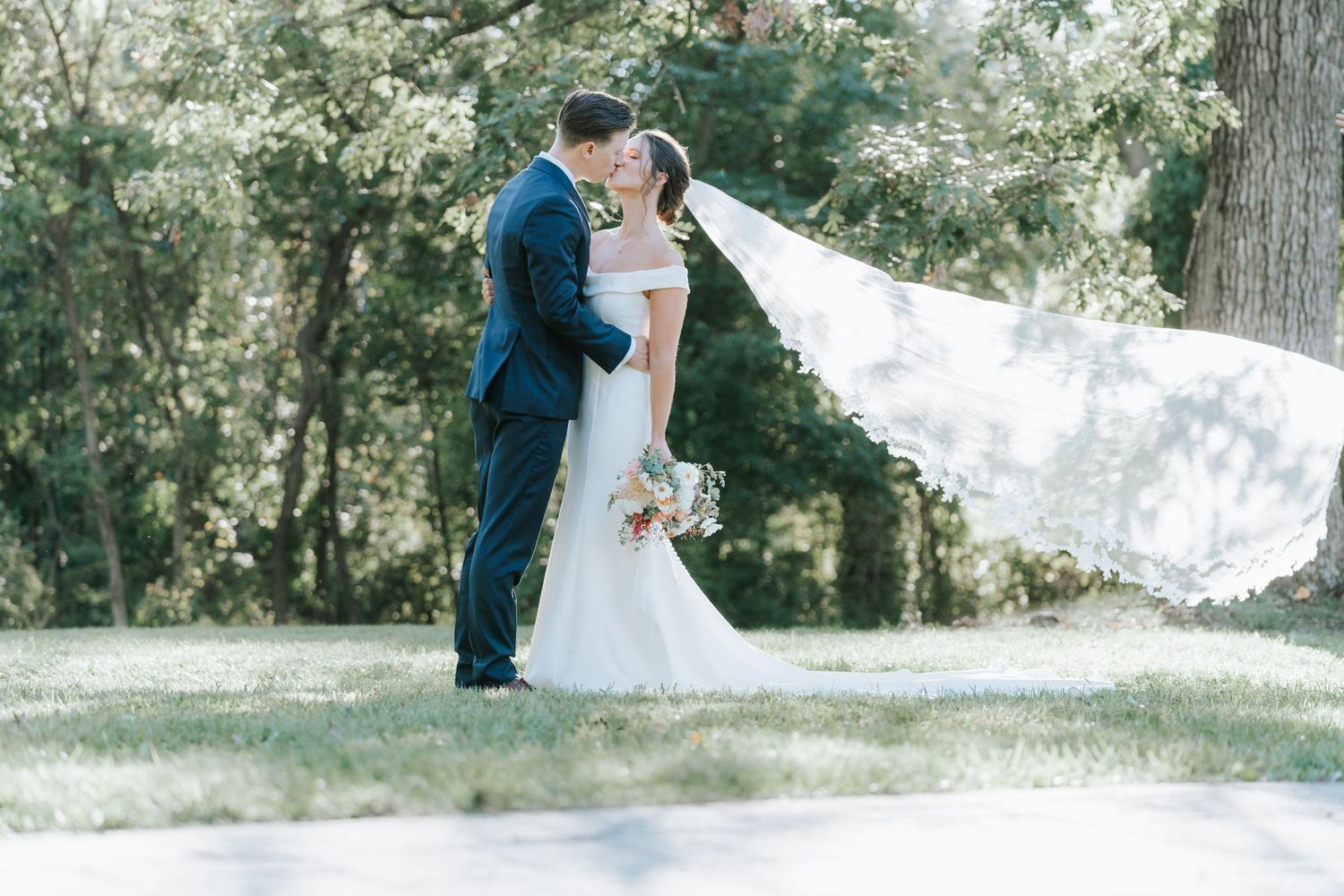 Dramatic wedding portrait with flowing veil in sunlit garden setting.