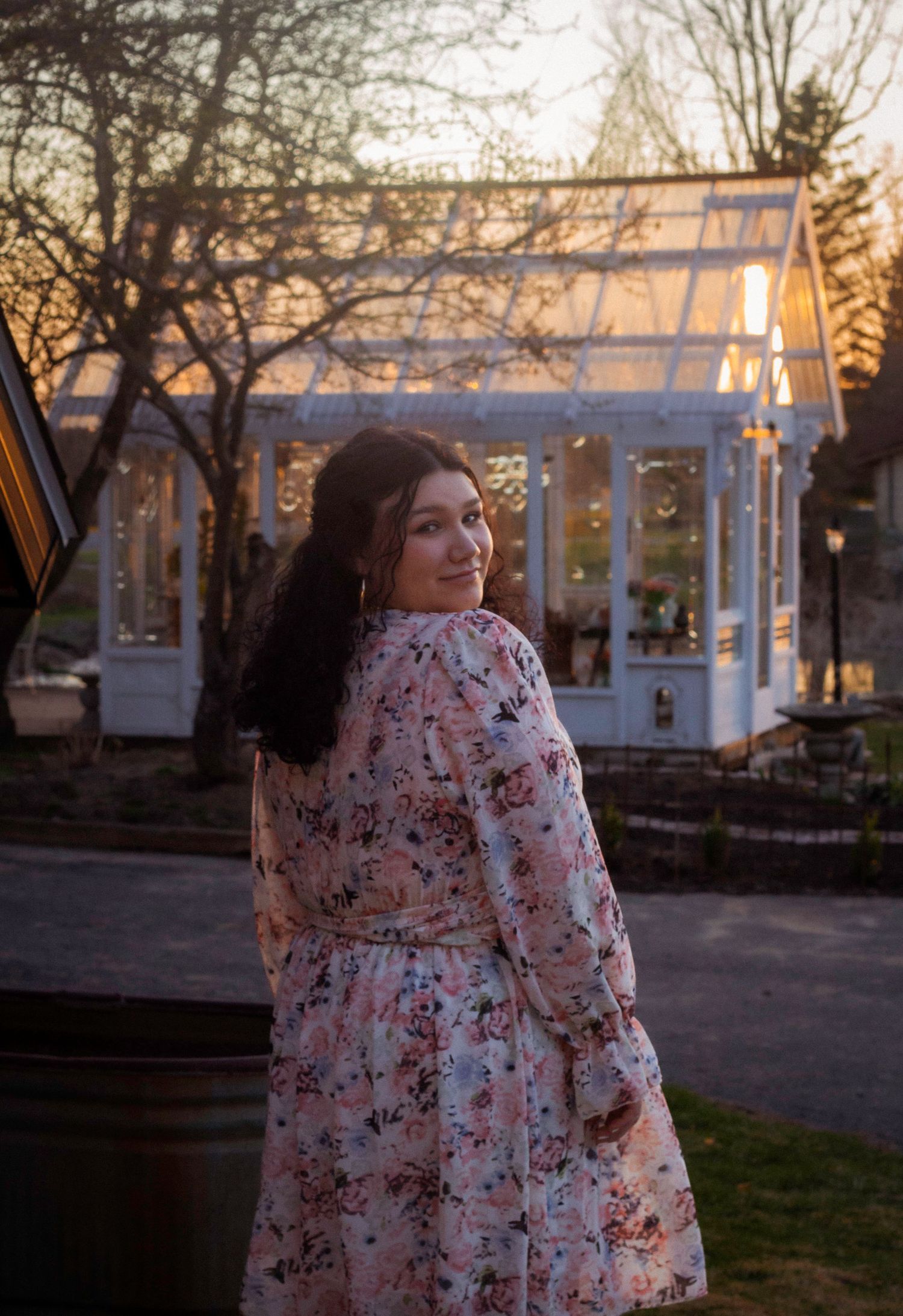 Person in a floral dress poses outdoors at sunset near a glass conservatory building.