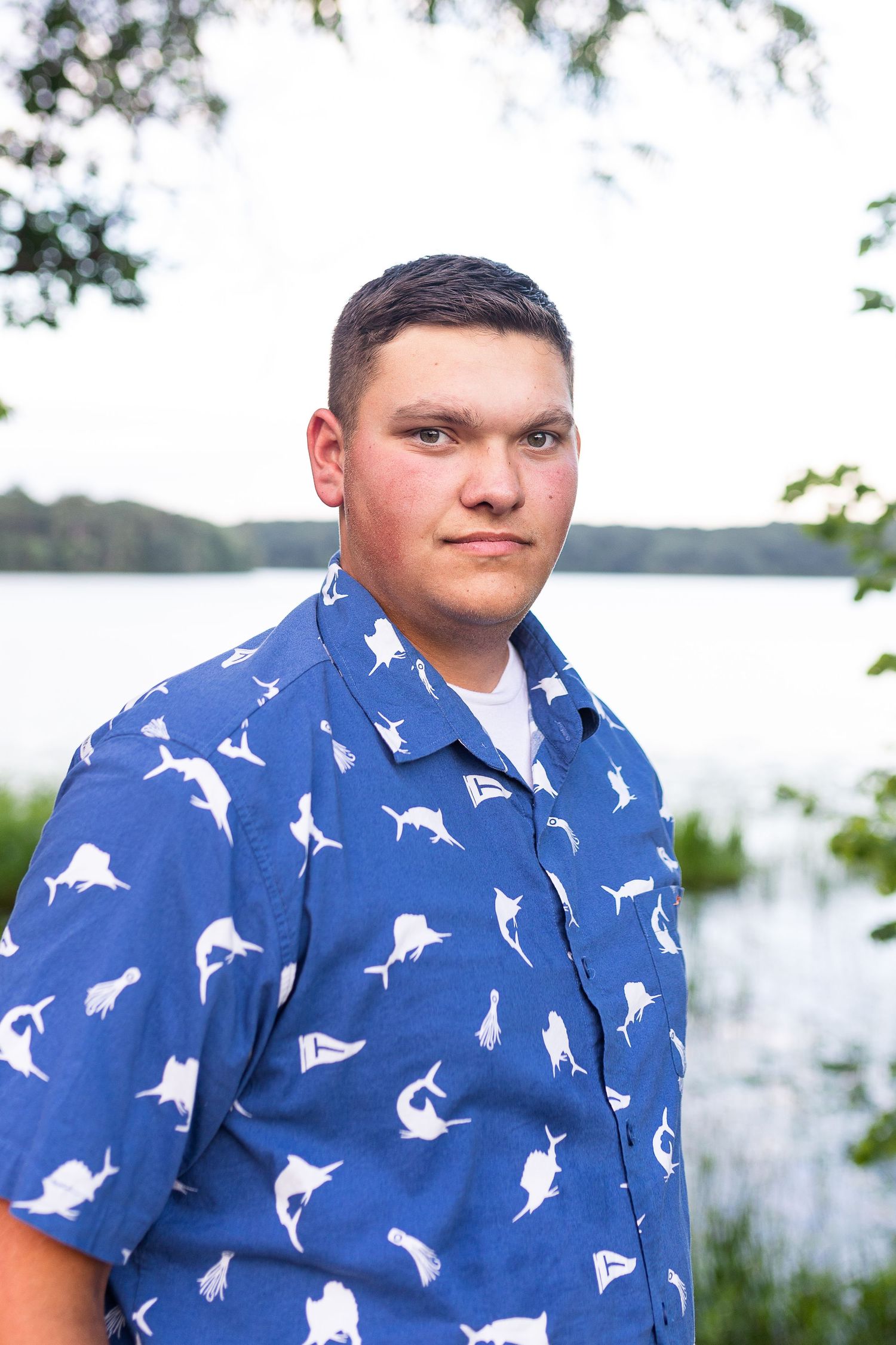 A person wearing a blue shirt with white dolphin prints poses by a lake.