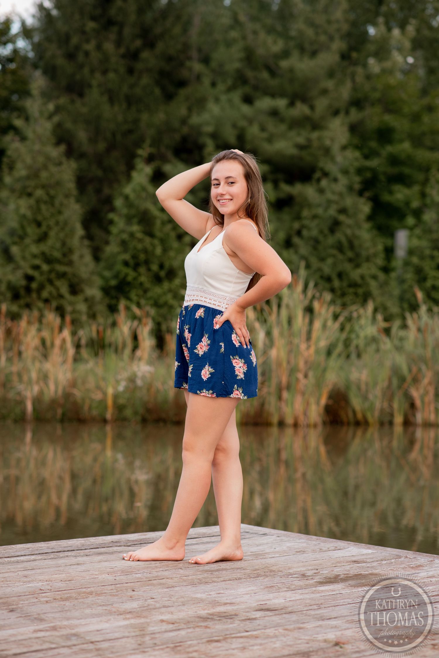 Full-length outdoor portrait of someone posing on a dock wearing a white tank top and floral shorts.